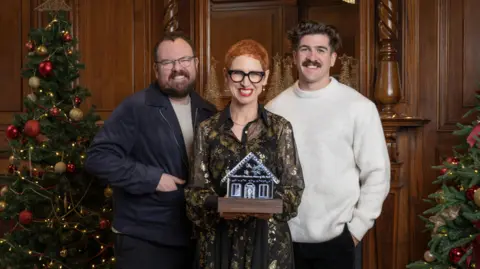 IWC The three judges of Scottish Home of the Year, Anna Campbell-Jones, Banjo Beale and Danny Campbell, stand smiling at the camera. Anna, who is in the middle, is holding the Scottish Christmas Home of the Year trophy, which is shaped like a gingerbread house. 