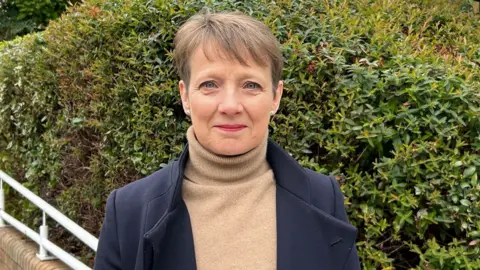 Clare Moody, wearing a brown top and navy jacket, looks ahead and smiles. She has short brown hair
