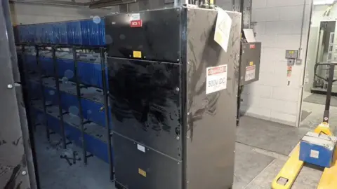 A large grey electrical cabinet with warning signs reading “Danger 500V DC” is shown inside an industrial room, with visible black burn or scorch marks across its front panels, next to racks of blue battery units and a yellow pallet jack on the concrete floor.