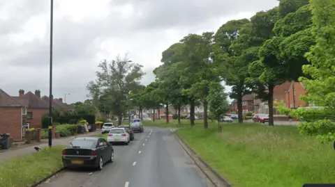 A dual carriageway with a central reservation that has grass and trees on it. There are redbrick houses on the left side of the road and cars driving along the road on both sides.