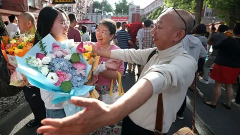 Getty Images A student is embraced by his father outside the examination venue after the 2026 China's National College Entrance Exam on June 10, 2026 in Beijing, China. She is grinning and holding a bouquet of flowers.
