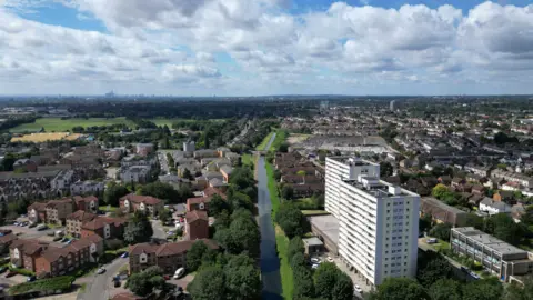 Getty Images Aerial view of a suburban area with a river running through the centre, surrounded by rows of houses, green spaces, and a tall residential tower block in the foreground, under a partly cloudy sky.