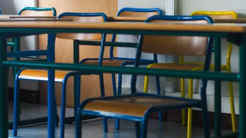 Wooden chairs on desks in a classroom. The chairs have green, blue and yellow metal.