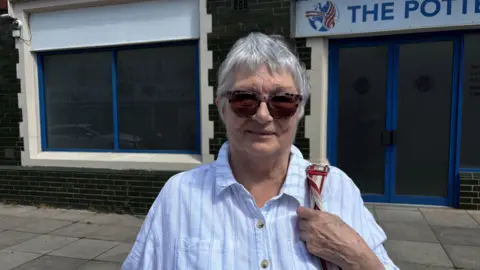 Gemma Dillon A lady wearing a blue striped shirt is pictured holding a bag.
She has short grey hair and sun glasses. 
She is stood in front of a closed building.