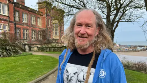 Michelle Lyons/BBC A middle-aged man with long flowing fair hair, which is partially plaited. He is wearing a blue jacket and a t-shirt with pictures of seagulls on it. Scarborough Beach is behind him.
