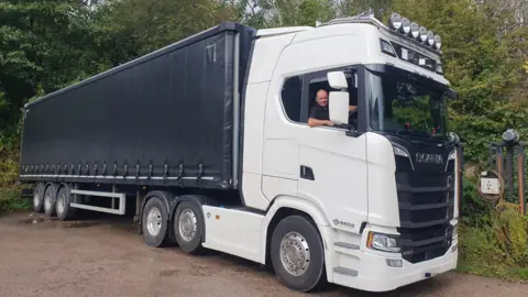 Michael Fletcher sitting in the driver's cabin of his lorry, with his right arm resting on the window ledge. It is a large transport lorry with a white cab and a black plastic canvas covering the goods.