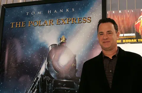 Getty Images Tom Hanks, wearing a black jacket and dark, striped shirt, stands in front of a promotional poster bearing his name and the title "The Polar Express". It features the front of a steam locomotive with a bell and and a bright light, set against a snowy sky. 