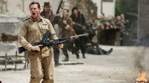 Banijay Rights/BBC Jack O'Connell, as Paddy Mayne in the TV drama, is seen shouting as he carries a Bren gun. In the background are flames and people running.