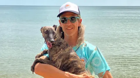 Carl Barnes stands looking at the camera, holding Margo the whippet and their glass trophy in his arms. They are standing with the sea in the background. Carl has shoulder-length light blonde hair. He wears a navy blue cap with a white panel on the front, which has the Shaka Surf logo on it. He is wearing pale blue round-framed sunglasses and a short sleeved pale blue t-shirt, which has the Shaka Surf logo on. Margo is perched over Carl's right arm and looks slightly off-camera, with her mouth open and tongue sticking out. She has a brindle coat, meaning her fur is a mixture of brown and black 'tiger stripes'.