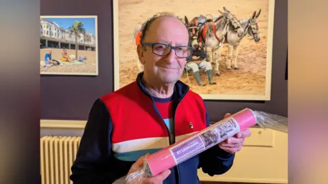Photographer JJ Waller at his Beside the Sea photography exhibition at the Hastings Museum and Art Gallery. JJ is wearing a striped Fila jacket and is holding a circular tube which says "Greetings from JJ Waller's St Leonards".
