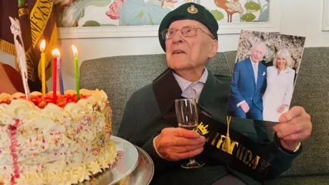Army veteran Sandy Calder wearing a green beret, holding a glass of fizz, and a birthday card featuring the King and Queen, as a birthday cake with lit candles arrived.