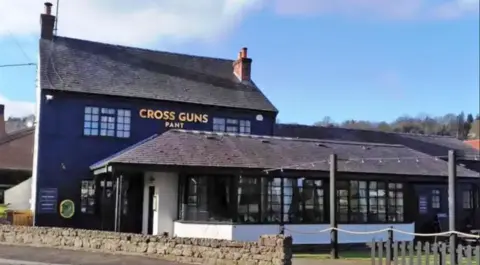 A two-storey pub with dark walls and a sloped roof with a one-storey extension at the front, also with dark walls and a sloped roof