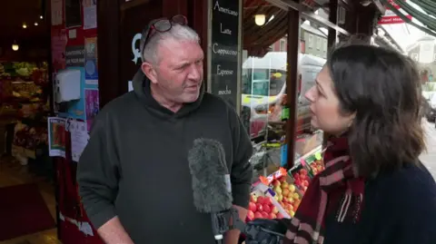 A man with grey hair and wearing a dark hoodie is being interviewed by a woman with brown hair outside a shop