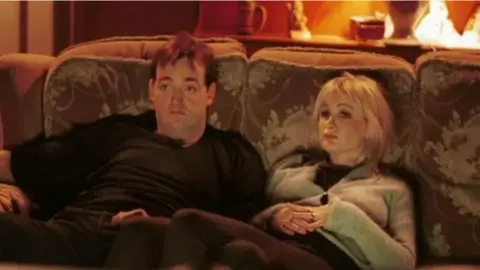 Craig Cash and Caroline Aherne playing Dave and Denise in the Royle Family. They are sitting on a sofa watching the television. 