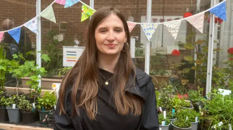 A woman with long brown hair wearing a black T-shirt with black waterproof jacket over the top. She is standing in front of a greenhouse with patterned bunting hanging on it. There are potted plants behind her. She also has a small gold necklace and gold hooped earrings.
