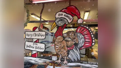 Debbie Carr The front of a butchers' windows with a painting on it. The painting shows a brown turkey with red tail feather and a red neck, wearing a red and white santa hat that reads "bah humbug". next to him is a red and white candy cane with signs on that read "merry christmas" and "from Battam's butchery"