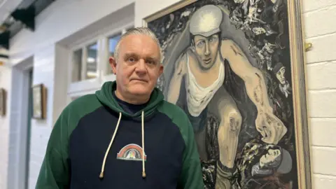 Man with brushed back grey hair, wearing a hoody with dark green hood and arms and black front, with a logo pit-head winding gear, stands in a white painted brick corridor, next to a portrait of a miner down in a pit 