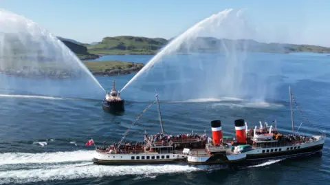 An mage of the Waverley paddle steamer making her way along a sunny coast, with a boat shooting water cannons into the air from either side as she passes.