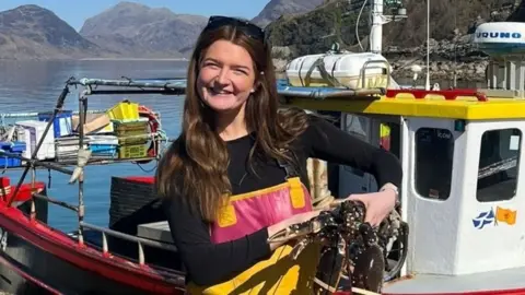 Brooke Lamond holds a lobster in front of a small creel fishing boat. She wears a long-sleeve black top with yellow fishing overalls on top. It is a sunny day and you can see the water behind her and hills in the distance.