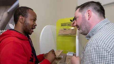 North Yorkshire Council The co-chairman of Grow Scarborough, Darren Mancrief, is pictured in front of a desk containing seeds in brown packets. He is showing these seeds to a visitor Athenkosi Nyengane