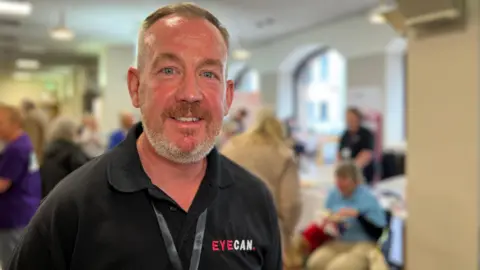 Mark smiles at the camera as he wears a black polo t-shirt with the EYECAN logo on it. Behind hime - people are talking to each other in the background. Mark has short grey hair and a short white beard.