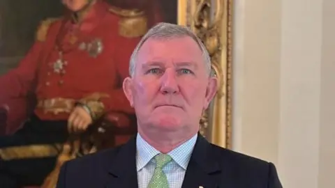 Richard Cripwell is standing in front of a historic portrait. He is wearing a dark coloured suit with a blue checked shirt and a green patterned tie. He is also wearing a military pin badge.