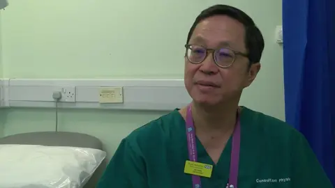 Dr Alan Tang, a man wearing teal medical scrubs with a purple lanyard and green NHS badge. Behind him is a hospital bed with a pillow on it, and a blue medical curtain.