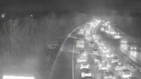 A traffic camera image above the M11 motorway. It is night time and the vehicles on both sides of the carriageway have their lights on. The carriageway closest to the camera has long queues of traffic. 