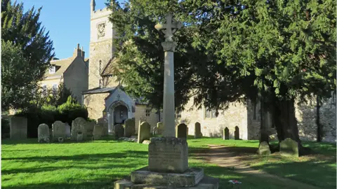 John Sutton/Geograph Grey stone war memorial in churchyard, surrounded by lawn, gravestones, a large tree and church in background 