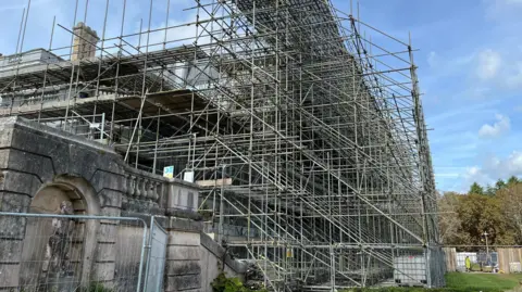 Torbay Council The outside of the mansion which is completely covered in scaffolding. The building is barely visible behind the scaffolding. There is fencing around it and a construction site in the background.