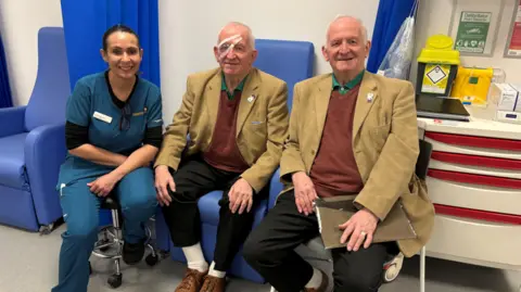 The twins are wearing brown jacket, red jumpers and green shirts with black trousers, white socks and brown shoes. They are sitting in blue hospital style chairs with a woman posing next to them. The brother in the middle has a clear patch and tape over his right eye. 