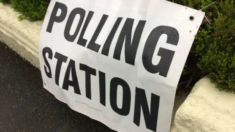 A canvas sign saying "Polling Station" attached to a low wall and a hedge.