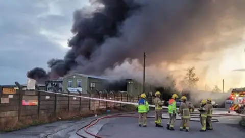 A group of firefighters in brown uniforms with yellow helmets stand near a concrete fence at the side of an industrial estate, where a large plume of black and grey smoke rises from green buildings