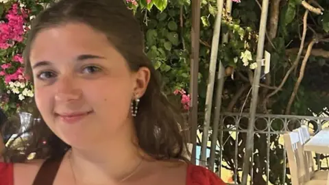 Chloe Hurst A woman in a red dress looks at the camera in front of a green bush and pink flowers. 