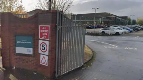 A view of Cheswold Park Hospital from its gates. The hospital name is on a sign on a wall and there is a car park with a building at the rear