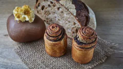 Simpsons Break that has been rolled up into a spiral and baked standing on a woven mat with a loaf that has been cut in half behind it.