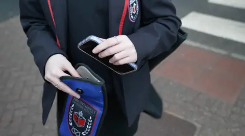 Close-up of a pupil in a dark school blazer with red trim placing a smartphone into a small blue pouch bearing the school crest, standing on a pavement.