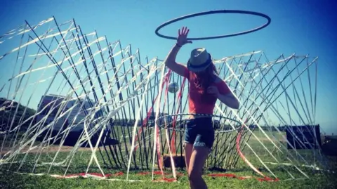 Lowri Denma Lowri dancing with a hula hoop around her waist. She is wearing a red short-sleeved top and denim shorts and in a field with an art installation covered in streamers behind her.