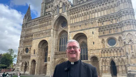 A man with a black blazer and clerical collar has black rimmed glasses on, grey hair and is smiling into the camera. Behind him is the front of a grand cathedral building.