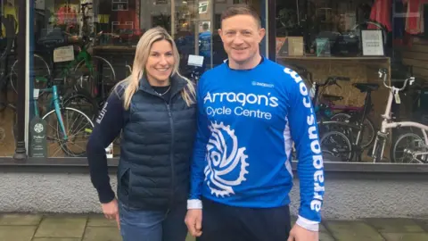 Arragon's Cycles Sarah and Phil standing in the foreground. Sarah has long blonde hair and Phil is wearing a blue T-shirt which says Arragon's Cycle Centre on it. Behind there is a window with lots of bikes inside it. The couple are smiling.