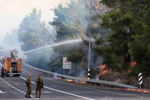 Getty Images Israeli firemen spray water on trees that are burning by the side of a road