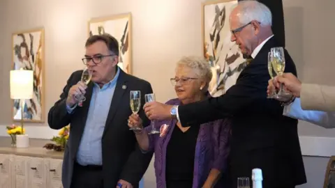 Amira Choice and Ebenezer Senior Living Anita LeBrun wears a purple blazer and glasses as she holds up a flute glass of champagne to toast Minnesota Gov Tim Walz, who is taller, balding and holding his own glass close to hers