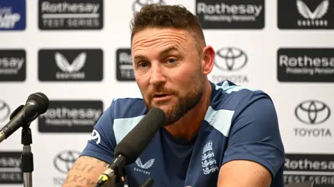 Brendon McCullum speaks into a microphone with sponsor's boards behind