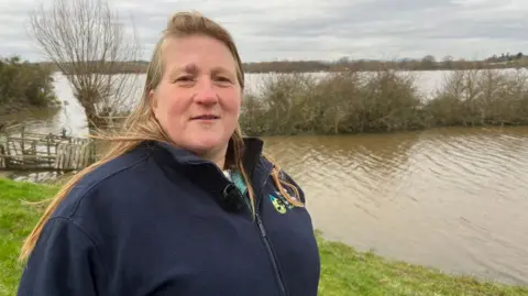 Debbie Wilkins stands by a flooded field on her farm on a winter's day. She has long blonde hair and wears a navy-blue fleece.