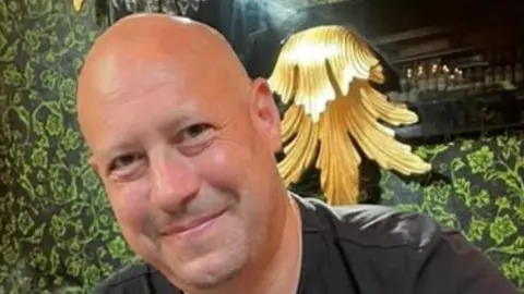 Hertfordshire Constabulary/Contributed Olly Frogell: A head and shoulders image of a bald man who is leaning to the right and smiling. He is wearing a black, round-necked top. Behind him is a green patterned wall and what appears to be a gold light fitting. 