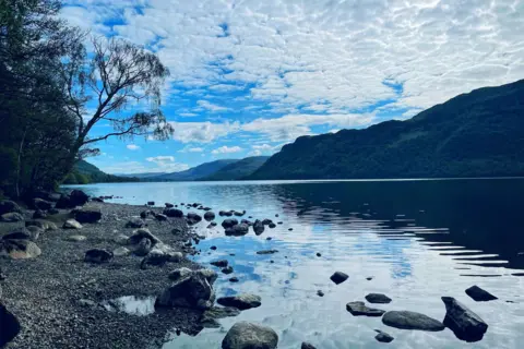 Georgia Roper A view of the lake from a rocky shoreline where some of the larger rocks, which are placed further out, rise above the water. The large mountain in the distance is reflected in the water below. The blue of the sky is barely visible through the white clouds.