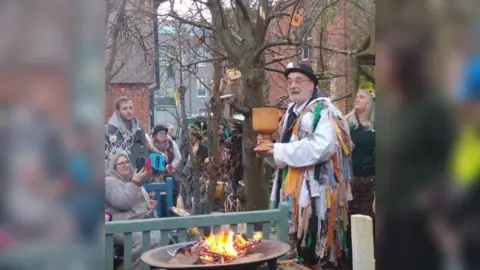 The Folk of Gloucester Bill wearing his 'tatter' coat, which is covered in long ribbons of varied colours. He is holding a large goblet, with a fire in front and a tree behind, which has pieces of toast hanging from the branches. A crowd is surrounding the tree.