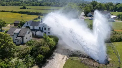 PA Media A aerial shot of the house damaged by the burst water pipe in Matson, taken from a drone. A huge plume of water is visible cascading over the house