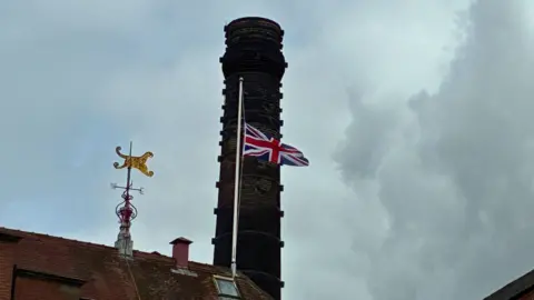 BBC/Fiona Callow A Union Jack flag on top of a brewery flies at half mast against a cloudy grey sky.