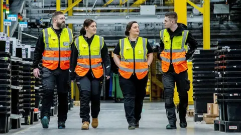 Four people in hi-viz vests and dark clothing are walking abreast through a warehouse. Two women are in the middle of the line, with a man at either end.